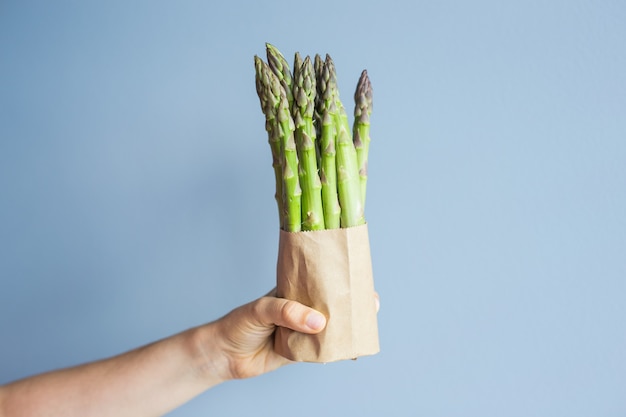 Female hand holds bundle of green asparagus on blue background concept of vegans vegetarians and