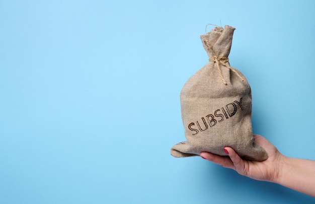 Photo a female hand holds a burlap sack with the inscription subsidy on a blue background concept of increasing government subsidies to the population