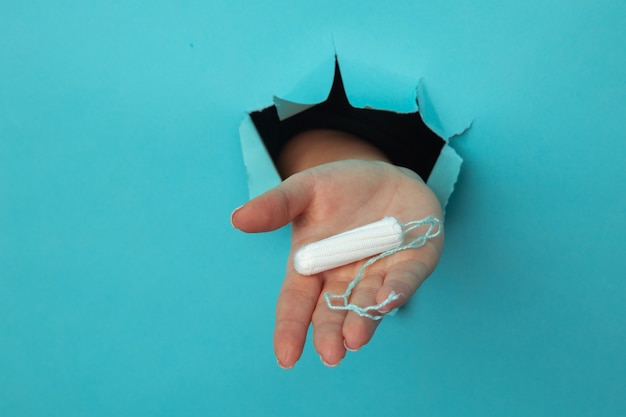 Female hand holds cotton tampon through torn paper background, health care concept