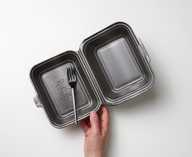 Photo female hand holds a disposable food container and a plastic fork. gray blank polystyrene box on white background