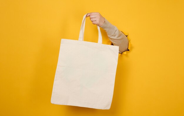 Female hand holds an empty white textile bag on a yellow background, a part of the body sticks out of a torn hole in a paper background. Reusable and recyclable packaging, no plastic