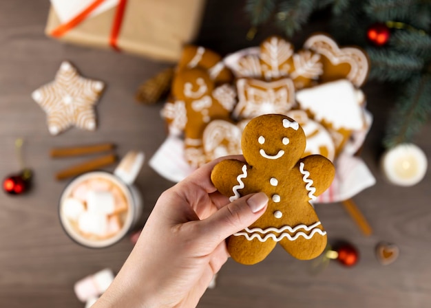 A female hand holds a gingerbread man on a background of a gingerbread basket a cup of cocoa with marshmallows a gift box and Christmas tree branches