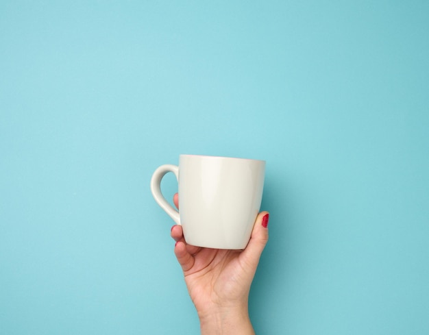 Female hand holds a gray ceramic mug on a blue background, break time and drink coffee