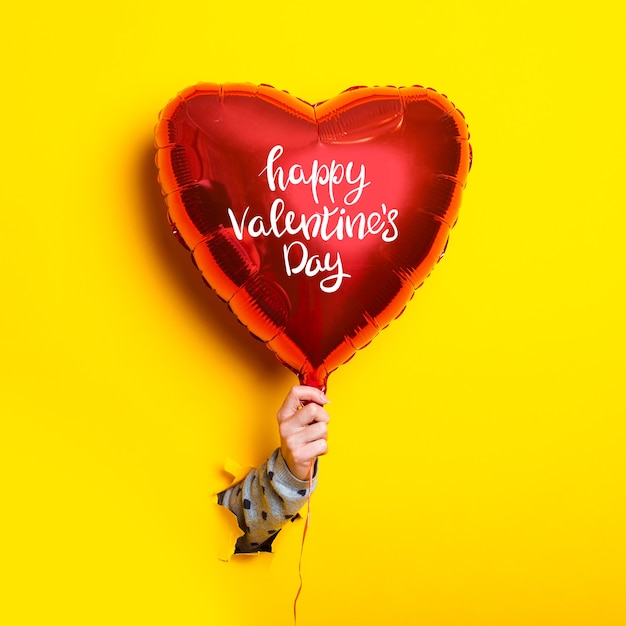 Female hand holds an inflatable balloon with the inscription Happy Valentines Day on a torn yellow background.