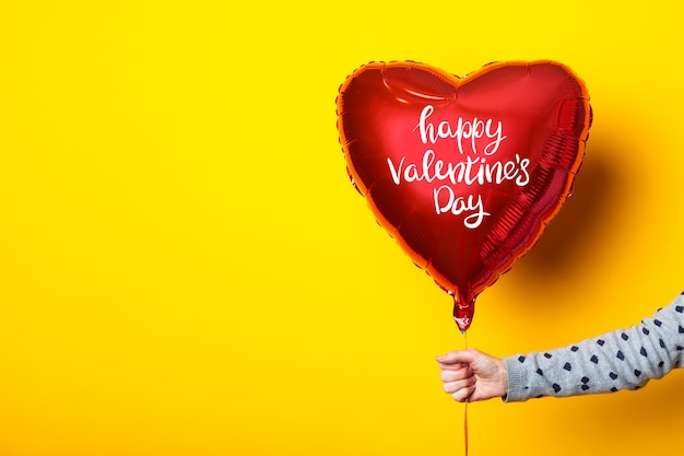 Female hand holds an inflatable heart-shaped balloon with the inscription Happy Valentines Day on a yellow background.