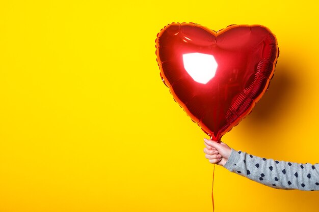 Female hand holds an inflatable heart-shaped balloon on a yellow background.