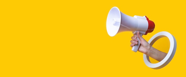 Female hand holds a megaphone in a round hole on a yellow background. banner