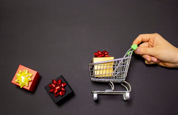 A female hand holds a metal trolley with a box on a black background with empty space for text. Black Friday sale.