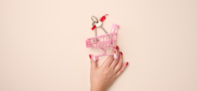 Female hand holds a miniature metal shopping cart with keys on a beige background. real estate purchase, mortgage, discount, banner