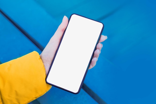 Female hand holds a mobile phone with a blank white screen on a blue wooden background Mockup