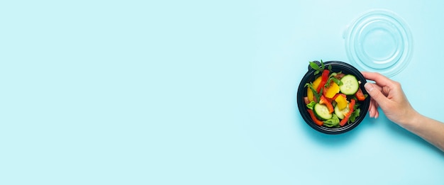 A female hand holds an open plastic disposable plate with vegetable salad on a light blue background. Top view, flat lay. Banner.