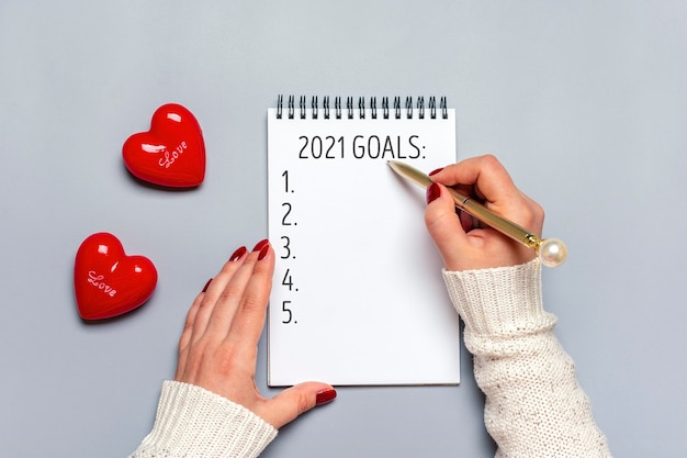 사진 여성 손에 펜을 들고 흰색 메모장에 2021 새해 목표를 기록하고 회색에 두 개의 빨간색 하트를 씁니다.