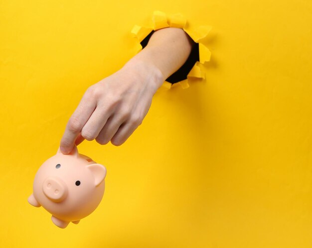 Female hand holds piggy bank through torn hole yellow paper Concept art Minimalism