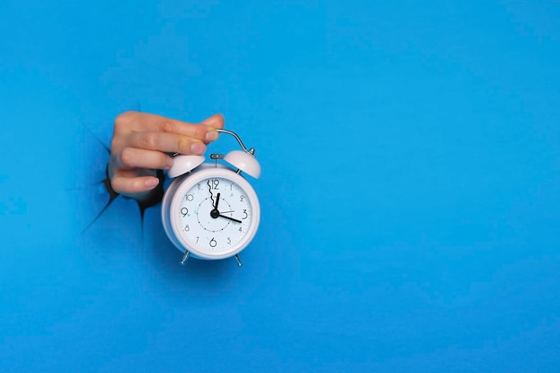 Female hand holds pink alarm clock through a paper hole in blue background