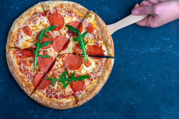 Female hand holds pizza with culinary shovel. Fresh prepared pepperoni pizza with salami and cheese on a dark background. Italian traditional lunch or dinner. Fast food and street food concept.