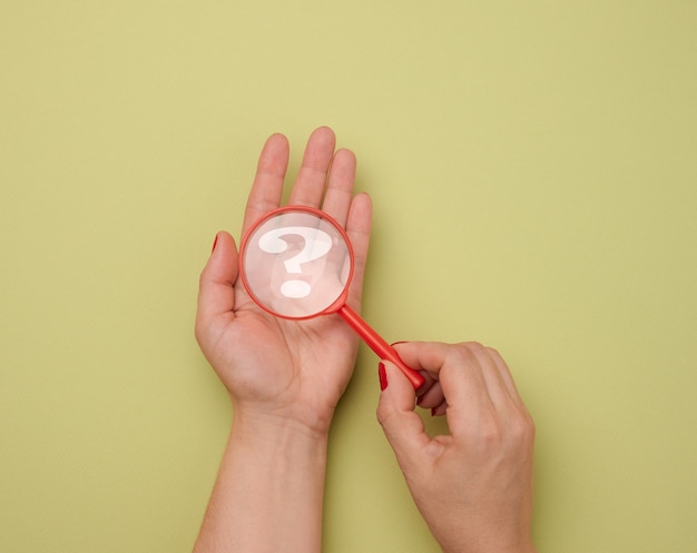 女性の手は、緑色の背景にプラスチックの虫眼鏡と疑問符を持っています。質問、真実、不確実性に対する答えを見つけるという概念。