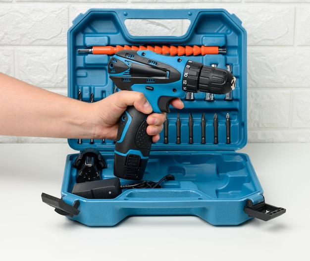 A female hand holds a portable drill on a battery against the background of an open plastic suitcase with attachments