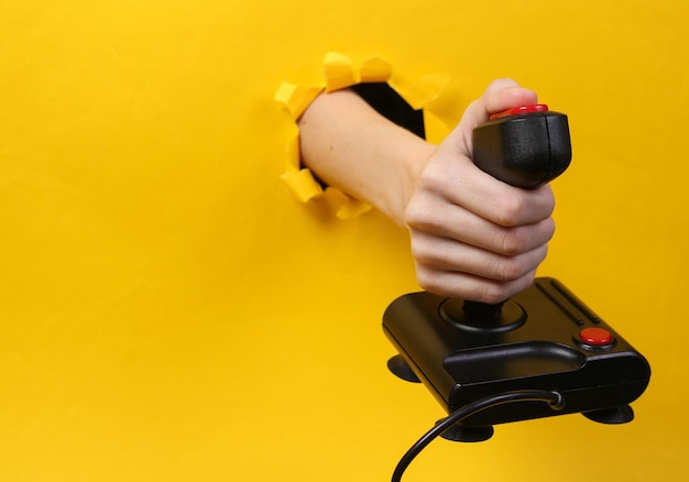 Female hand holds retro joystick through torn hole yellow paper Concept art