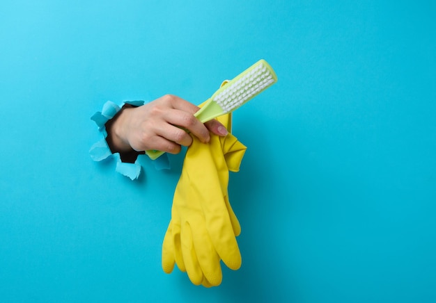 Female hand holds rubber gloves and a plastic cleaning brush, part of the body sticks out of a torn hole in a blue paper background