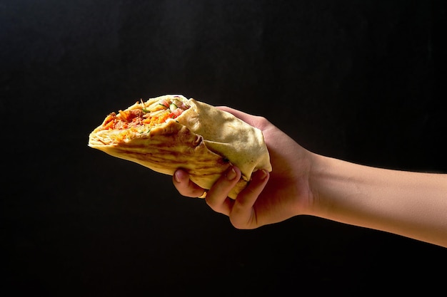 Female hand holds shawarma with red sauce, meat and vegetables on a dark background