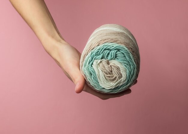 Female hand holds a skein of woolen threads on pink pastel studio background.