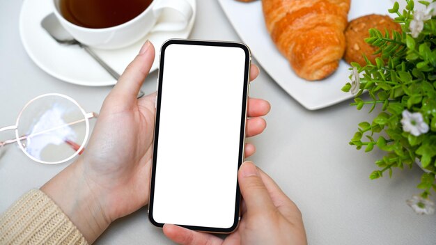 Female hand holds a smartphone white screen mockup over a coffee table top view