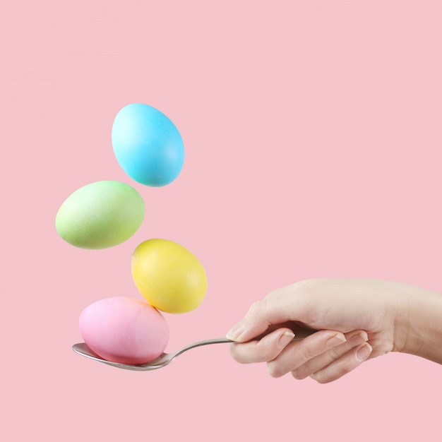 Female hand holds a spoon on which multi-colored eggs are balanced, on a pink background. Unusual design, Easter concept, copy space.