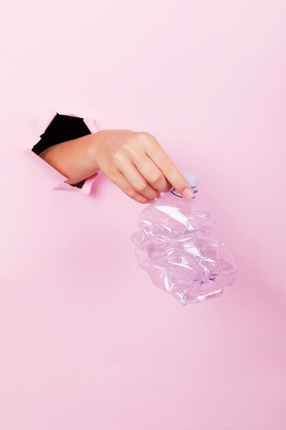 Female hand holds through a hole a empty crumpled plastic bottle on a pink background, no plastic and zero waste concept