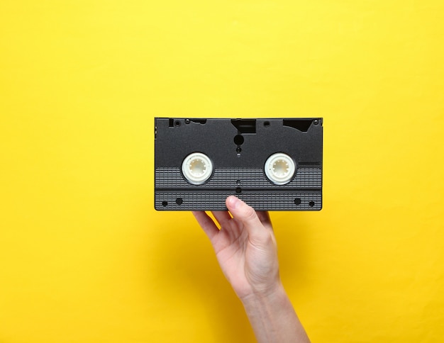 Female hand holds video cassette on yellow background. Retro style, pop culture, minimalism, top view