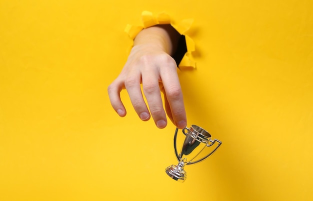 Photo female hand holds winner cup through torn hole yellow paper concept art minimalism