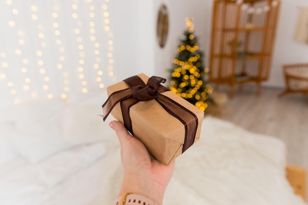 Female hand is holding a decorated gift blurred Christmas tree wrapped present celebrating and giving conceptChristmas zero waste Gift box packed in paper decorated with lace and twine