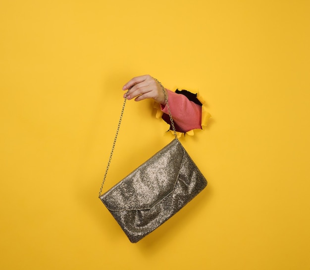 Female hand is holding a small golden clutch bag with a metal chain on a yellow background. Part of the body sticking out of a torn hole in a paper background