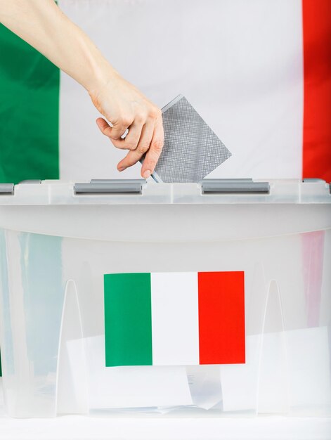 女性の手が投票箱の上に投票を保持します。背景のイタリア国旗。閉じる