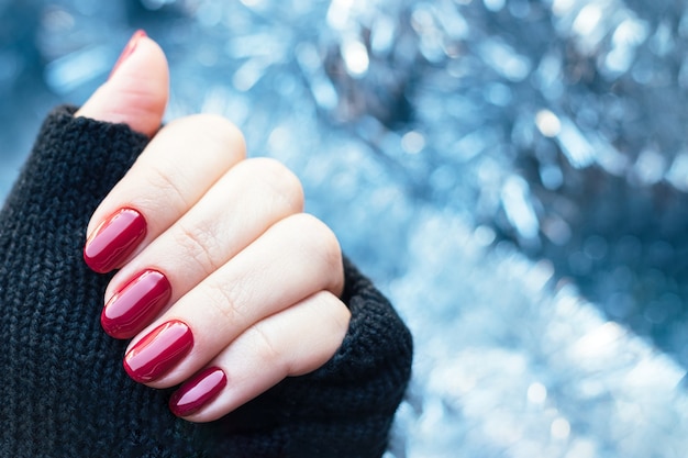 Female hand in knitted gloves mitts with glossy dark red nails on background of Christmas tinsel