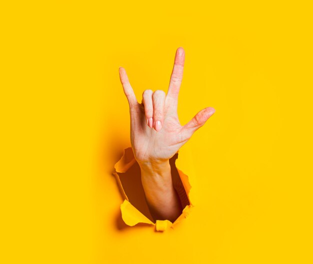 Female hand makes rock and roll gesture through hole in yellow paper Girl demonstrates horn sign Body language concept Hand in rock sign cool symbol