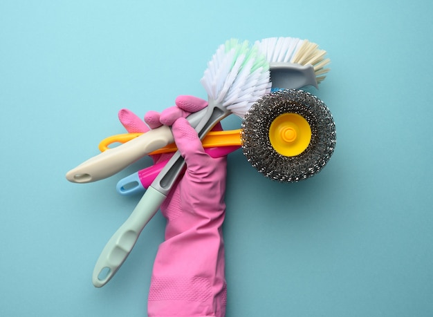 Female hand in a pink glove holds a stack of plastic cleaning brushes, blue background