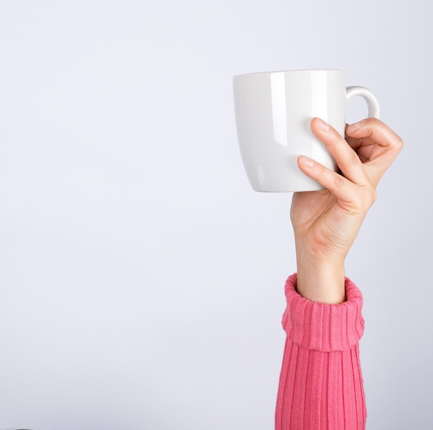 Photo female hand in a pink sweater holds a gray ceramic cup, white surface