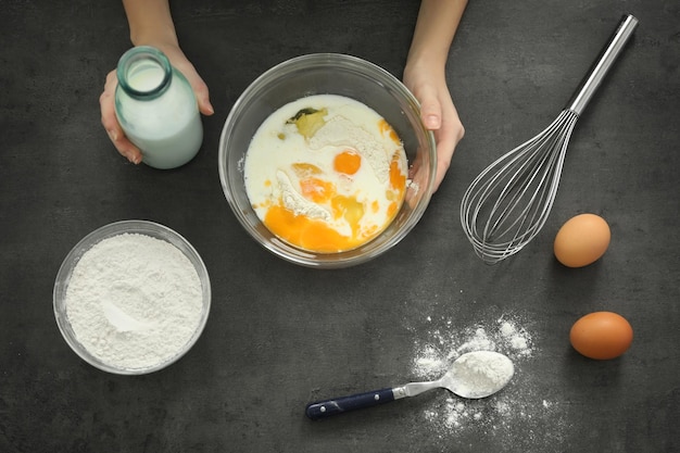 Female hand pouring milk into glass bowl with dough on table top view