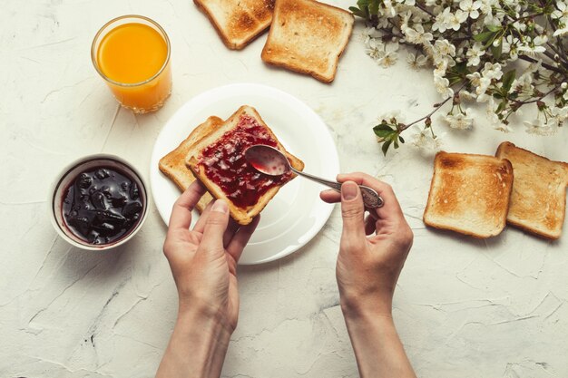 Female hand put jam on bread toast, glass of orange juice, sprigs spring tree with flowers, white stone surface. breakfast concept. flat lay, top view