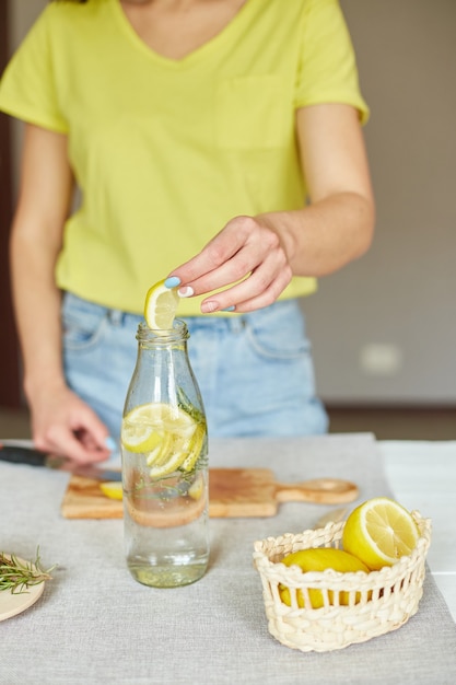 Female hand put lemon in glass bottle, Woman preparing, making detox healthy water with lemon and rosemary, fresh lemonade in glass on a white table at home, summer drink