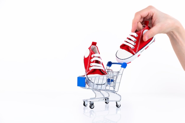 Female hand puts baby red sneakers into the shopping cart isolated on white background. Online store of children's shoes. Copy space.