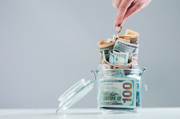 Female hand puts a coin into a glass piggy bank full of money.