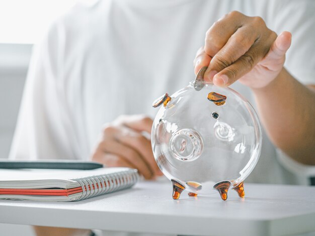 Female hand putting coin into piggy bank on white table closeup