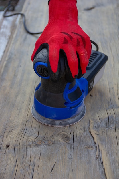 Female hand in red protective glove is grinding an old gray board with an electric sander, front view with copy space