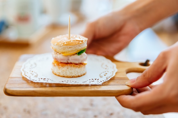 Female hand serving Mini Chicken Burger on wooden chopping board with blur background.