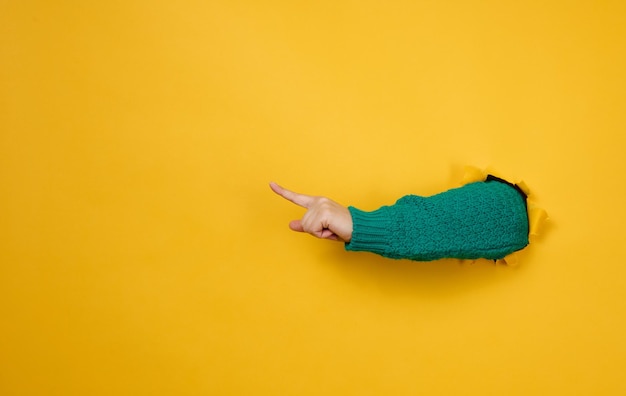 Female hand sticks out of a torn hole in a yellow paper background part of the body points with the index finger to the side copy space