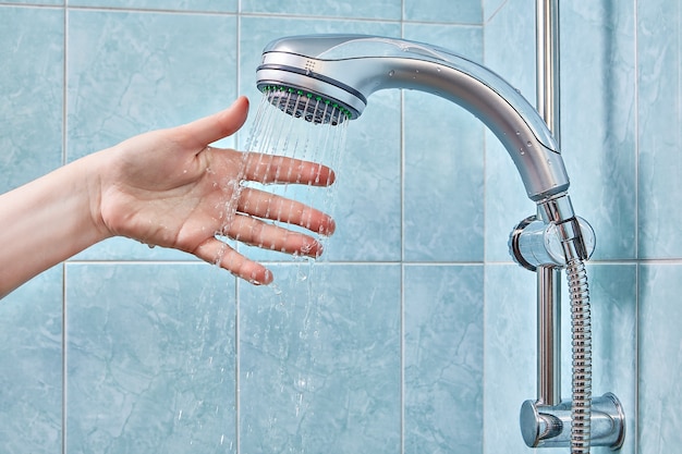 Female hand under stream of water from shower head, fixed in holder, checks water temperature in bathroom with blue tiles.