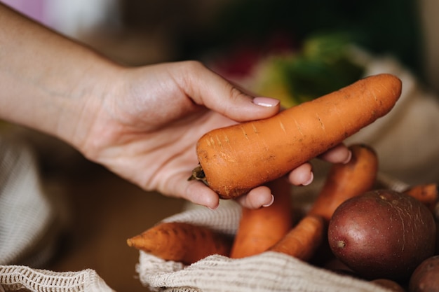 Женская рука берет морковь из холщового продуктового мешка. Овощи в многоразовых мешках из эко-хлопка на деревянном столе. Концепция покупок с нулевыми отходами. Товары без пластика