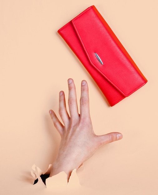 女性の手は、引き裂かれた黄色のパステル紙を通して赤い財布を取ります。ミニマルなクリエイティブなファッションコンセプト