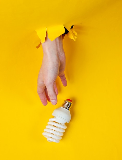 Photo female hand takes spiral light bulb through torn yellow paper. minimalistic creative fashion concept